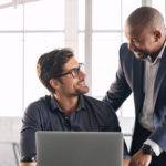 Senior and junior businessman discussing during their meeting. African american executive working with young businessman in modern office. Mature boss with business partner working together on laptop.